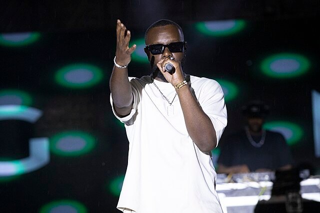 Le chanteur Gims en t-shirt blanc et lunettes de soleil, chantant dans un micro sur scène lors d'un concert.