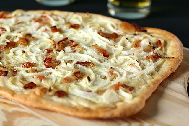 Photo d'une tarte flambée alsacienne traditionnelle (Flammekueche) dorée et croustillante sur une planche en bois, illustrant la gastronomie locale et les mots alsaciens du terroir.
